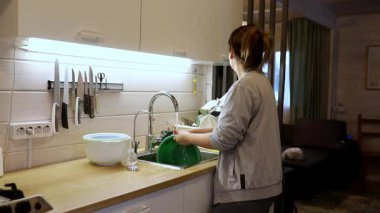 Bir kadın iyi aydınlatılmış bir mutfakta bulaşıkları yıkıyor. Mutfak aletleri ve aletleriyle çevriliyken yeşil bir kase kullanıyor. Güneş ışığı pencerelerden geçer, sıcak atmosferi güçlendirir..