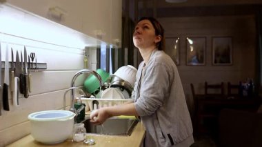 Bir kadın sıcak, davetkar bir mutfakta lavaboda duruyor, akşamları bulaşıkları yıkıyor. Doğal ışık, tabakları temizlerken odaklanmış yüz ifadesini aydınlatır..