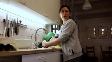 Bir kadın sıcak ve davetkar bir mutfakta lavaboda duruyor, yemekten sonra bulaşıkları yıkıyor. Yumuşak ışık, günlük angarya işleri yaparken sakin bir atmosfer yaratıyor..