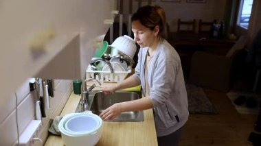 Çağdaş bir mutfakta bulaşıkları yıkayan bir kadın. Etrafı temiz çatal bıçaklarla çevrili. Mekan aydınlık ve rahat, öğleden sonra sıcak bir ev atmosferini yansıtıyor..