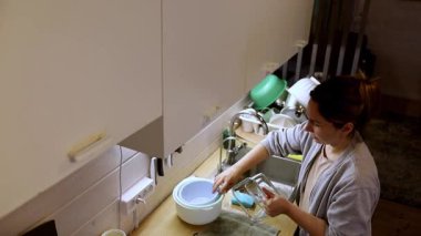 Bir kadın mutfak lavabosunda duruyor, bulaşıkları sabunlu suyla yıkıyor. Güneş ışığı pencereden içeri akar, temiz ve düzenli alanı vurgular..