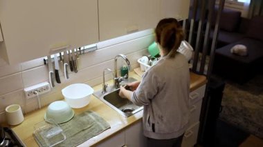 Bir insan, doğal ışıkla dolu parlak bir mutfakta bulaşıkları yıkamakla meşgul. Mekanda modern cihazlar ve rahat bir atmosfer var..