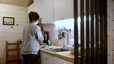 Bir kadın sıcak ve davetkar bir mutfakta bulaşıkları yıkıyor, ahşap aksanlar ve etkin ışıklandırmalarla, öğleden sonra rahatlatıcı bir atmosfer yaratıyor..