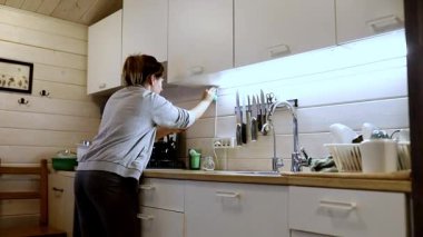 Bir kadın kırsal bir kulübede mutfağını topluyor, bıçakları düzenliyor ve tezgahı temizliyor. Doğal ışık boşluğu doldurur, sıcak atmosferi güçlendirir..