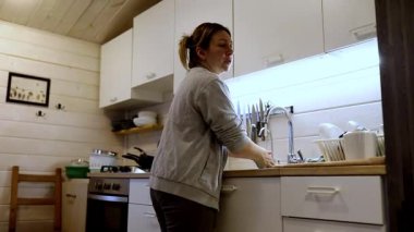 Ahşap aksanlı ve modern aletli rahat bir mutfakta bir kadın yemekten sonra bulaşıkları yıkıyor. Yumuşak ışık, mutfak alanını temizlerken sıcak bir atmosfer yaratıyor..
