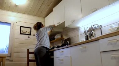 Bir kadın büyüleyici ahşap bir kulübede mutfak dolaplarını temizleyip düzenliyor. Sıcak öğleden sonra ışığı, çeşitli mutfak malzemelerini toparlarken gösterdiği çabayı vurguluyor..