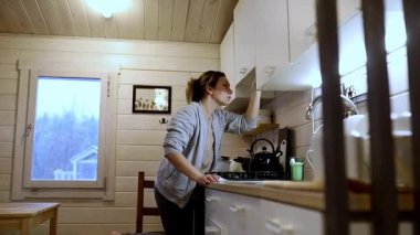 Bir kadın yağmurlu bir öğleden sonra büyüleyici bir ahşap kulübede mutfağını topluyor. Raftaki eşyalara uzanıp sıcak ve davetkar bir atmosfer yaratıyor..