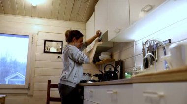 Büyüleyici ahşap bir kulübede mutfak dolaplarını düzenlemek için uzanan bir kadın görülüyor. Sıcak atmosfer ve yumuşak ışık huzurlu bir akşam ortamı yaratır..