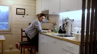Bir kadın bir sandalyede otururken rahat bir kulübede mutfağı temizliyor. Yumuşak doğal ışık boşluğu dolduruyor, ahşap dekorun altını çiziyor. Sahne rahat bir ev atmosferi yakalıyor..