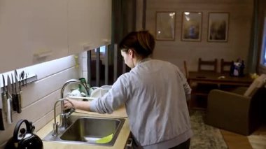 Bir kadın mutfağını toplar, bulaşıkları yıkar ve güneş ışığı odayı doldururken düzenler. Mekan sıcak ve davetkar. Sakin bir öğleden sonra atmosferi var..