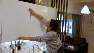 Bir kadın çağdaş bir evde mutfak dolaplarını özenle temizler. Her köşesinin lekesiz olduğundan emin olmak için bir bezle uzanıyor..