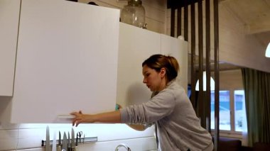 Bir kadın, çağdaş bir evde mutfak dolaplarını iyice temizlerken görülüyor. Mutfak sıcakken toprağı temizlemeye odaklanmış. Hoş bir atmosfer yaratıyor..