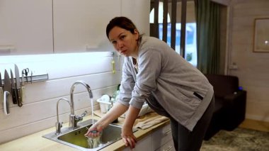 Bir kadın mutfak lavabosunu süngerle temizlemekle meşgul. Tertipli, davetkar bir mutfakta. Atmosfer sıcak, doğal ışık sıcak atmosferi güçlendiriyor..