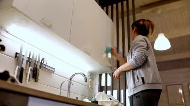 Bir kadın şık, modern bir mutfakta mutfak dolaplarını düzenleyip temizliyor. Etrafı çatal bıçaklarla çevriliyken yüzeyleri siliyor, temizlik ve düzeni vurguluyor..