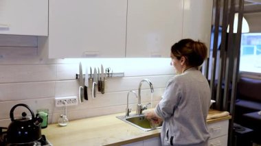 Zarif, modern bir mutfakta, bir kadın titizlikle lavaboda bulaşıkları yıkarken etrafı düzenli çatal bıçaklar ve bolca ışıklandırmayla çevrili, temiz ve verimli bir yer sergiliyor..