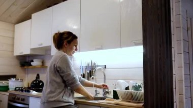 Hoş geldin kabin mutfağında, bir kadın bulaşıkları lavaboda yıkayarak ev işleriyle ilgilenirken doğal ışık içeri süzülüp huzurlu bir atmosfer yaratıyor..