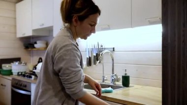 Bir kadın mutfak lavabosunun önünde durmuş sıcak ve büyüleyici bir alanda bulaşıkları yıkıyor. Güneş ışığı pencereden süzülerek modern dolap ve huzurlu atmosferi vurguluyor..