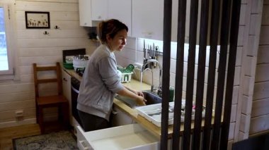 Parlak bir mutfakta, bir kadın lavaboda bulaşıkları yıkamakla meşgul. Sabah ışığı odayı aydınlatıyor ve sıcak bir atmosfer yaratıyor. Mutfak derli toplu, minimalist dekorları var..