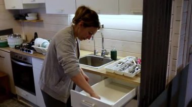 Bir kadın, modern bir evde mutfak aletleri ve mutfak aletleriyle dolu bir mutfak çekmecesini karıştırırken görülüyor. Yemek yapmak için etrafı toplarken odaklanmış görünüyor..