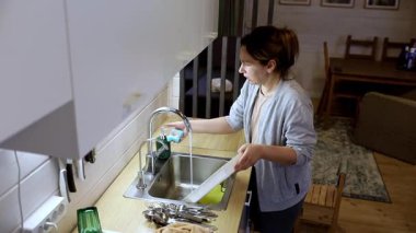 Bir kadın sıcak, davetkar bir mutfakta bulaşıkları yıkıyor, çatalları dikkatlice düzenliyor ve lavabonun düzenli kalmasını sağlıyor. Atmosfer rahat ve sade..