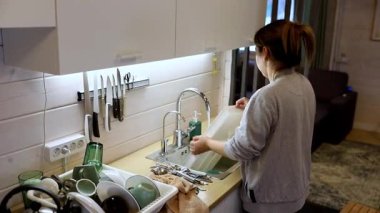 Parlak bir mutfakta bulaşıkları yıkayan bir kadın. Etrafı temiz çatal bıçaklar ve camlarla çevrili. Doğal ışık boşluğu doldurur ve sıcak bir atmosfer yaratır..