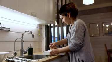 Mutfak lavabosunda bulaşık yıkayan bir kadın, sıcak, davetkar, yumuşak ışıklandırma ve ahşap aksanlarla dolu bir yerde gününü yansıtıyor. Akşamın parlaklığı ortamı daha da güzelleştiriyor..