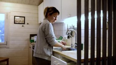 Bir kadın sıcak ve davetkar bir ahşap mutfakta bulaşık yıkıyor, yumuşak ışıkla aydınlatılıyor. Akşam çöktüğünde ortam sakin ve huzurlu..