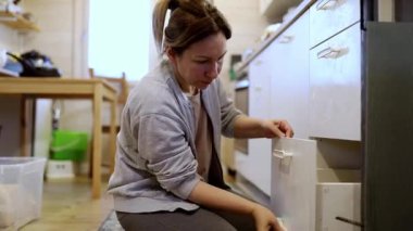 Bir kadın zamanını mutfak çekmecelerini düzenleyerek parlak ve davetkar bir mutfakta diz çökerek geçirir. Kolay erişim ve temizlik için düzenli olarak eşyalar düzenler..