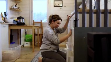 Bir kadın, kırsal bir kulübede kompakt bir mutfakta eşyalar düzenlerken görülüyor. Yerdeki konteynırları dikkatlice ayarlıyor, sıcak ve davetkar bir atmosferle çevrili..