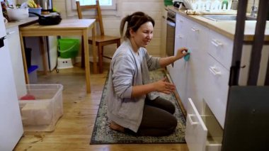 Bir kadın, küçük, rahat bir evde mutfak dolaplarını özenle temizliyor. Diz çöküp, öğleden sonra ışığında temiz bir yer tutma konusundaki kararlılığını gösteriyor..