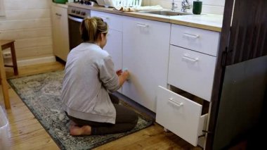 Davetkar bir mutfak ortamında, bir kadın alt dolapları düzenlemek için diz çöker. Doğal ışık pencereden süzülerek sıcak ve misafirperver bir atmosfer yaratır..