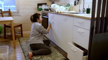 Bir kadın yerde diz çöküyor, dolapları özenle temizliyor ve parlak bir mutfak alanında düzenliyor. Tabaklar istiflenmiş, atmosfer hoş karşılanıyor ve düzenli..