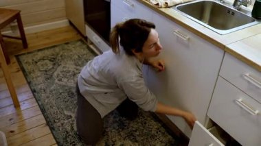 Bir insan sıcak bir mutfağın zemininde diz çöker, dolapların kapılarını süngerle ovar. Güneş ışığı içeri akar, temizlik ve düzenleme için hoş bir atmosfer yaratır..