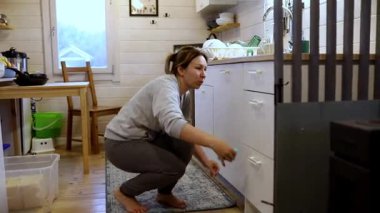 Bir kadın, rahat kır evinde mutfak dolaplarını düzenlerken çömelmiş olarak görülüyor. Güneş ışığı içeri akar, sıcak ve davetkar bir atmosfer yaratır..