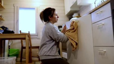 Bir kadın, rahat bir mutfakta diz çöküp, yüzeyleri temizliyor ve mutfak aletlerini düzenliyor. Güneş ışığı pencereden içeri akarak sıcak bir atmosferi vurgular..