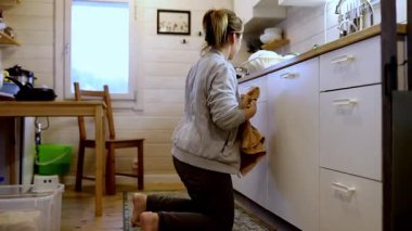 Bir kadın, aydınlık ve davetkar bir mutfakta dolap içeriği düzenliyor, yerde diz çökerken eşyaları dikkatlice yerleştiriyor, ev bakımı ve organizasyondaki çabasını gösteriyor.