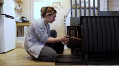 Bir kadın ocak içine dikkatlice bir kütük yerleştiriyor ve odadaki sıcaklığı arttırıyor. Ahşap iç mekan ve yumuşak ışıklandırma soğuk mevsimde rahat bir atmosfer yaratır..