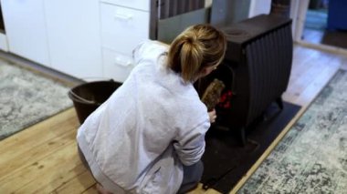 Bir kadın, odunları yakan bir sobaya eğilimli, şık yaşam alanında sıcaklık yaratmak için odunları dikkatlice ekliyor. Oda modern ve kırsal unsurların karışımından oluşuyor, rahatlığı artırıyor..