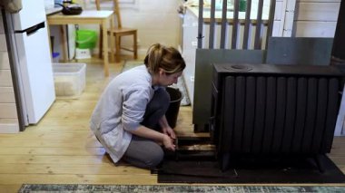 Rahat giyimli bir kadın, ahşap bir kulübede ısıtma sobasını ayarlıyor. Önümüzdeki kış ayları için sıcaklığı sağladığı için doğal ışık boşluğu dolduruyor..