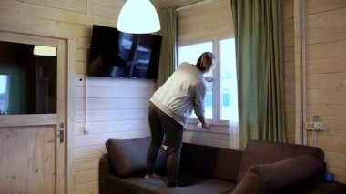 Bir kadın, yüksek noktalara ulaşmak için kanepenin üzerinde duran bir kulübenin camlarını temizler. Güneş ışığı içeri akar, doğal ahşap elementlerle dolu rahat iç kısımları vurgular..