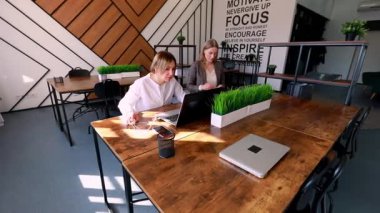 Motivasyon ve yaratıcılığı vurgulayan çağdaş bir ofis alanında dizüstü bilgisayarlar üzerinde çalışırken iki kadın tartışıyorlar. Dekoratif bitkiler çalışma alanını geliştirir.