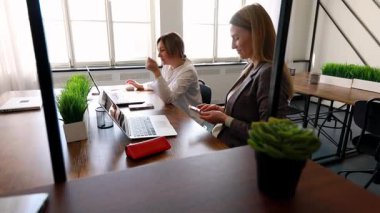 Çağdaş ofis ortamında çalışan iki kadın var. Biri dizüstü bilgisayar kullanıyor, diğeri de telefonunda. Atmosfer, konsantrasyon ve işbirliğini teşvik eder..