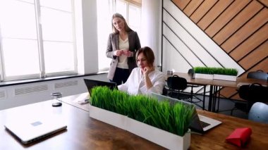 Çağdaş bir ofiste iki kadın bir tartışmaya girer. Biri diğerini bir dizüstü bilgisayarda çalışırken gözlemliyor. Ayarda doğal ışık ve bitki dekoru yer alıyor..