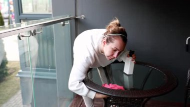 Evin içinde şık bir yerde bir kadın cam masayı dikkatlice temizliyor. Bir sprey şişesi kullanarak, temiz bir ortamda lekeleri temizlemeye odaklanıyor..