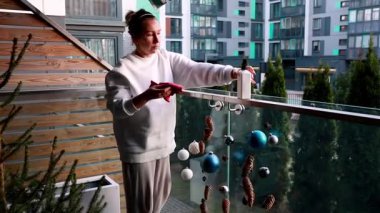 Bir kadın, modern bir apartman kompleksinde güneşli bir kış gününün tadını çıkarırken balkon korkuluklarına renkli süsler ve doğal elementler yerleştirir..