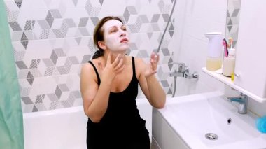 Banyosunda oturan bir kadın cilt bakımının bir parçası olarak yüz maskesi takıyor. Modern banyoda geometrik fayanslar ve sakin bir atmosfer var..