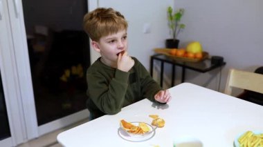 Genç bir çocuk sıcak bir mutfakta beyaz bir masada oturur ve narenciye dilimlerinin tadını çıkarır. Ortam sıcak ışık ve arka planda birkaç meyve ile dolu..