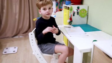 Genç bir çocuk renkli süslemelerle dolu bir odada masa başında oturuyor, yaratıcı oyunlarla ilgileniyor. Evdeki zamanının tadını çıkarırken kendini jestlerle çiziyor ve ifade ediyor..