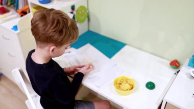 Parlak bir oyun odasındaki masada oturan genç bir çocuk patates cipsi yerken çizimlerine odaklanmış. Oda oyuncaklarla ve sanat malzemeleriyle dolu..