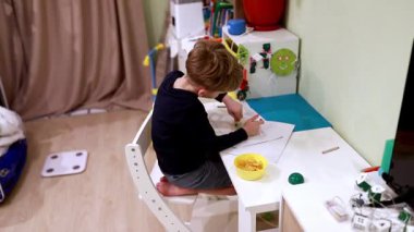 Küçük bir çocuk çizim etkinliğine odaklanmış küçük bir masada oturuyor. Oda sıcacık ve gevşemiş atmosferi arttırmak için yakınlara bir kase abur cubur yerleştirildi..